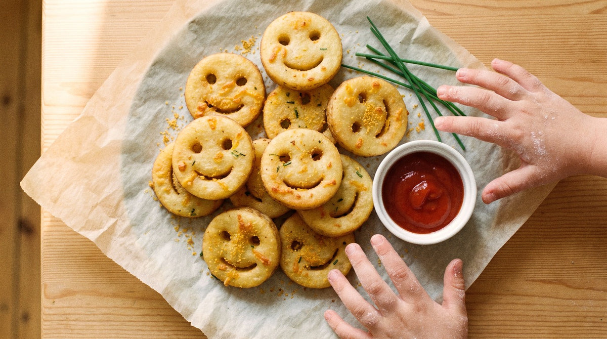 Homemade cheesy potato faces seasoned with Chooki's Original Chicken Salt on baking paper with tomato sauce and kids' hands reaching in