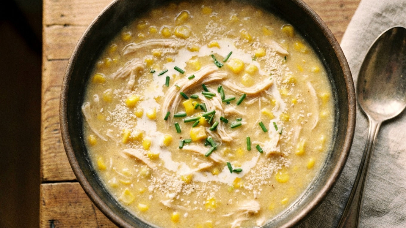A dark ceramic bowl of creamy chicken and sweetcorn soup seasoned with Chooki's Zest Chicken Salt, topped with cream and chives on a rustic wooden table