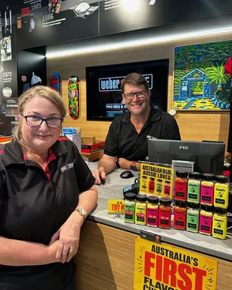 Two staff smiling at the counter of FNQ BBQ and Outdoors, beside a display of Chooki's chicken salt
